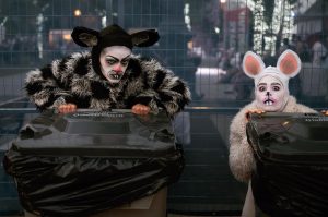 Two actors in rat costumes. One is black and grey and the other is white. They have large ears on their head, face paint, and fake teeth. 
They are sitting on garbage cans. One has a dark expression on their face and the other looks frightened. 