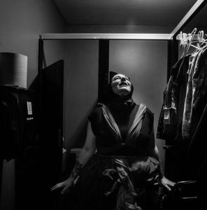 Promotional photo for middle child. A black and white photo of Kathleen Greenfield sitting on the ground looking up. She is wearing many layers and appears to have set pieces and clothing around her.