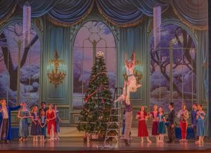 A Production photo of the Gift of the Nutcracker - Clara in a white dress is lifted up and is next to a large christmas tree. Several guests in beautiful clothing look up at her.