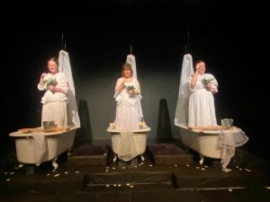 A production photo of drowning girls. They are dressed all in white and they are standing in claw footed bathtubs, holding a bouquet of flowers each.