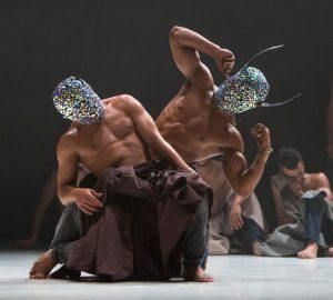 Dancers wearing sparkly masks and dark brown/black baggy trousers are in the centre of the image. One leans back and one poses while leaning to the side.