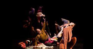 A promotional photo of the dance troupe. They are wrapped in layers of sweaters, and the dancer in the centre reaches up towards the light.