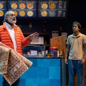 Two actors of Indian descent stand in a set that is made to look like a to go restaraunt. One wears a bright orange puffer coat and the other a brown polo tshirt. The one wearing a puffer coat holds a medium sized rug.