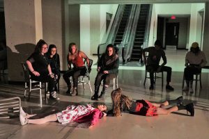 Performers in Concord Floral sit on chairs and one lays on the floor on their stomach. They are in an open, indoor space with an escalator behind them. 