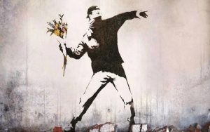 A famous image created by Banksy, a stencilled image on the wall. The image is of a man wearing a mask, winding his arm back as if ready to throw a bouquet of flowers. 