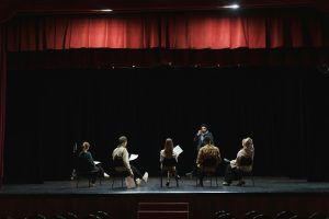 A stock image of a group of artists sitting on chairs on a stage, scripts in hand. 